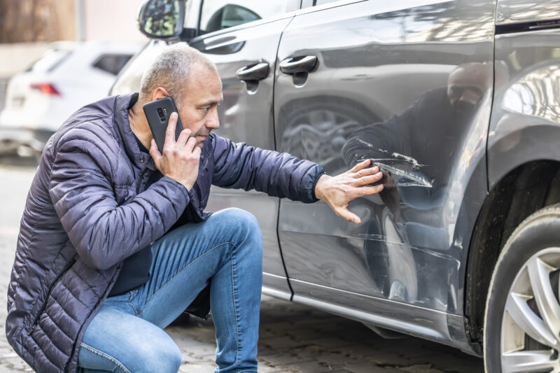 Man on his phone looking at accident damage to the door of his car