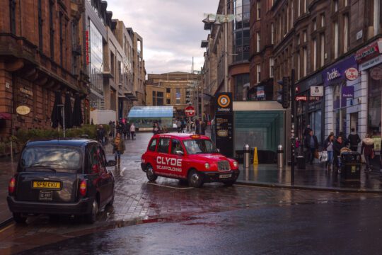 people walking down a high street in glasgow scotland next to a taxi rank with a red cab in the centre of the image