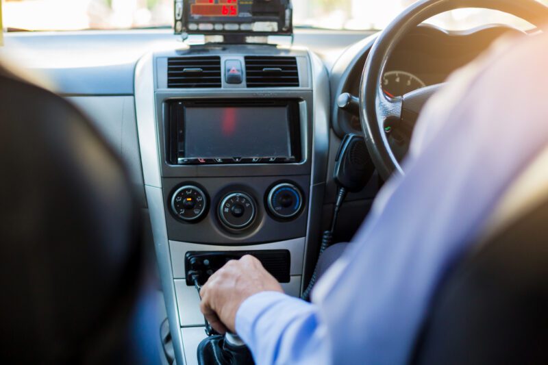 View of a taxi driver taken from behind. Taxi driver on the right side. Passenger seat is empty. The cars radio is in the centre of the image.