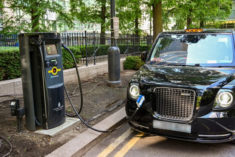 A black electric LEVC taxi is parked on a street while charging at a public charging station. The charging cable is plugged into the front of the taxi. The setting includes trees, a black metal fence, and modern glass buildings in the background.