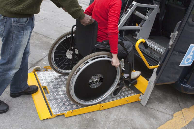 A person in a wheelchair wearing a red sweater and dark pants is being assisted onto a wheelchair-accessible taxi using a yellow metal ramp. A helper, wearing jeans and a green sweater, is standing nearby, holding the wheelchair for support. The taxi has a blue handicap-accessible sign on its door, and the interior features a securement system for the wheelchair. The scene takes place on a paved sidewalk with a yellow parking line visible on the ground.